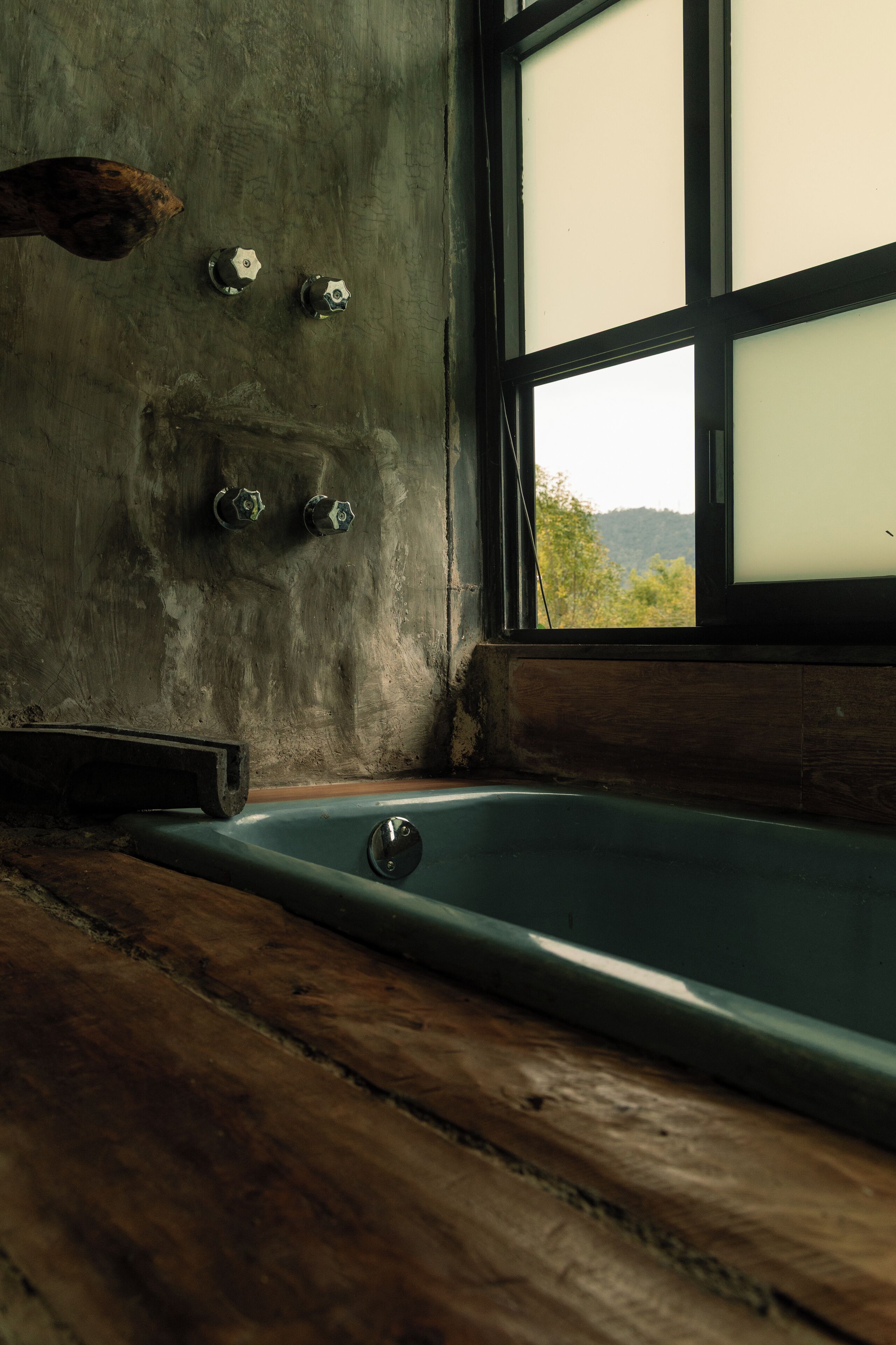 A bathtub in a rustic room with a large window, wooden floor, and a concrete wall with three faucets.
