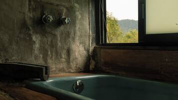 A bathtub in a rustic room with a large window, wooden floor, and a concrete wall with three faucets.