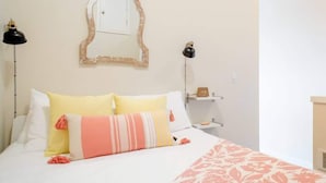 Una cama bien hecha con ropa de cama blanca y rosa, un espejo con marco decorativo y dos lámparas de noche.