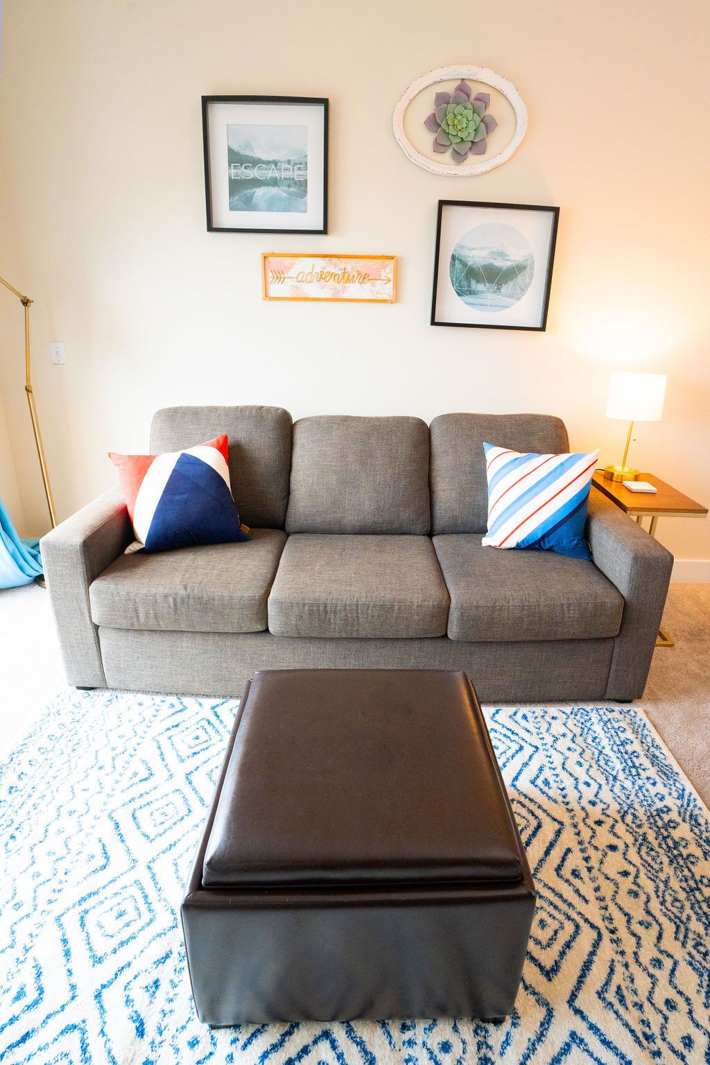 A living room with a grey sofa, two pillows, a framed picture, a small table with a lamp, and a patterned rug.