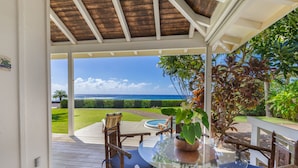 A covered outdoor area with a glass table and chairs, a view of the ocean, and a well-maintained lawn. - Modern Home with Hot Tub, AC & Ocean Views! (Po'ipu)