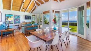 A spacious living area with a wooden ceiling, a large glass door leading to a view of the ocean, and a dining table with chairs. - Modern Home with Hot Tub, AC & Ocean Views! (Po'ipu)