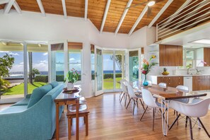 A modern kitchen with wooden cabinets, a dining area with a wooden table and white chairs, and large sliding glass doors leading to a view of the ocean.