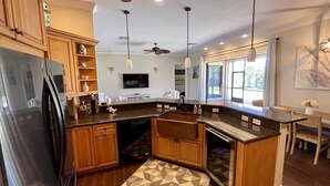 A modern kitchen with wooden cabinets, a black refrigerator, a dishwasher, a built-in oven, and a granite countertop. There is a ceiling fan, pendant lights, and a large window providing natural light. - Vacation Rental Home in Bonita Springs (Bonita Springs)