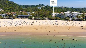 Una playa con numerosos paraguas y gente, un cartel que dice "ZINC NOOSA", y edificios al fondo.