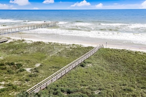 A beach with a wooden pier extending into the ocean, a grassy area with a fence, and a clear sky. - Top Floor Corner Unit | Beachfront Condo with Sweeping Ocean Views (Fernandina Beach)