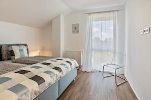 Un dormitorio con cama, una silla, un radiador y una ventana con cortinas.