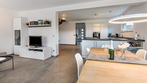 Una sala de estar moderna con televisor, mueble blanco para equipo multimedia y una zona de comedor con mesa y sillas de madera.