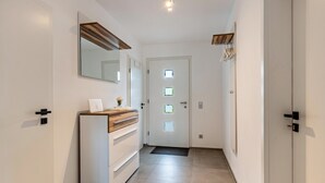 Un pasillo con una puerta blanca, un espejo, un mueble con cajones y un pequeño florero con flores.