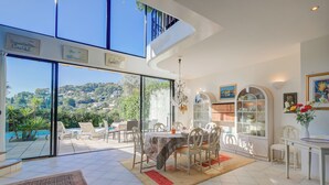 A spacious dining area with a table set for four, large windows offering a view of a hillside, and a glass-enclosed patio with outdoor furniture. - Semiramis Villa Laurie - Private pool, Mougins, France (Mougins)