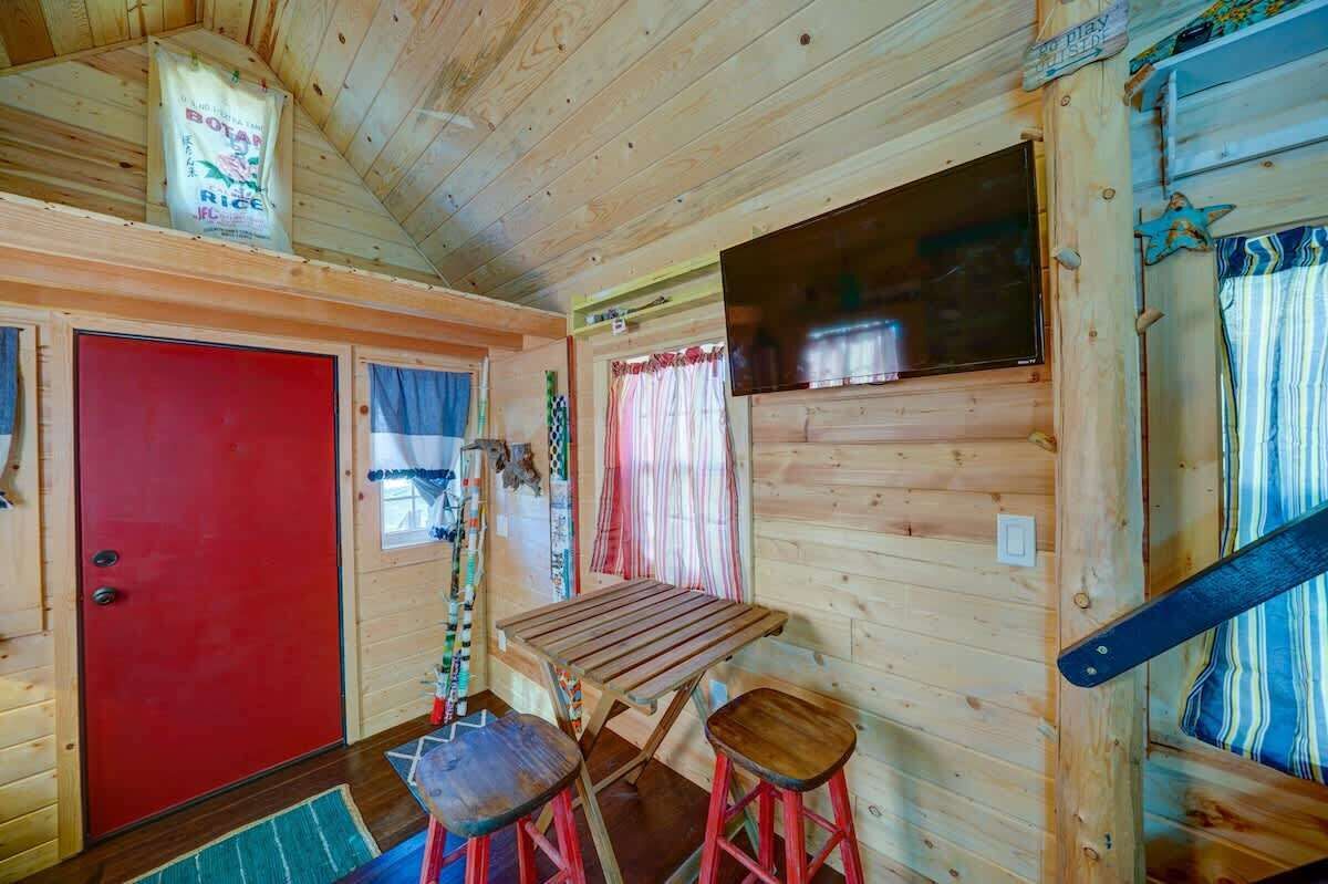 A wooden cabin interior with a red door, a small table, two stools, and a window with curtains.