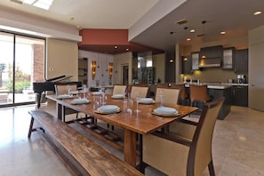 A spacious dining area with a long wooden table set for a meal, a piano, and a kitchen area with modern appliances.