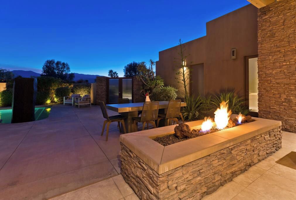A patio area with a stone fire feature, a dining table with chairs, and a pool in the background.