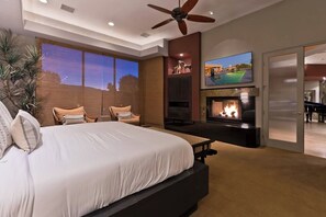 A bedroom with a large bed, a ceiling fan, a fireplace, a TV mounted on the wall, and a view of the outdoors through a window.
