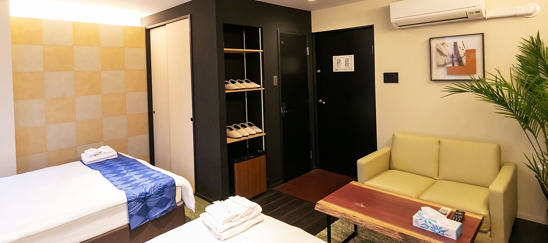 Quarto de hotel com cama, sofá, mesa pequena, sapateira e planta em vaso.