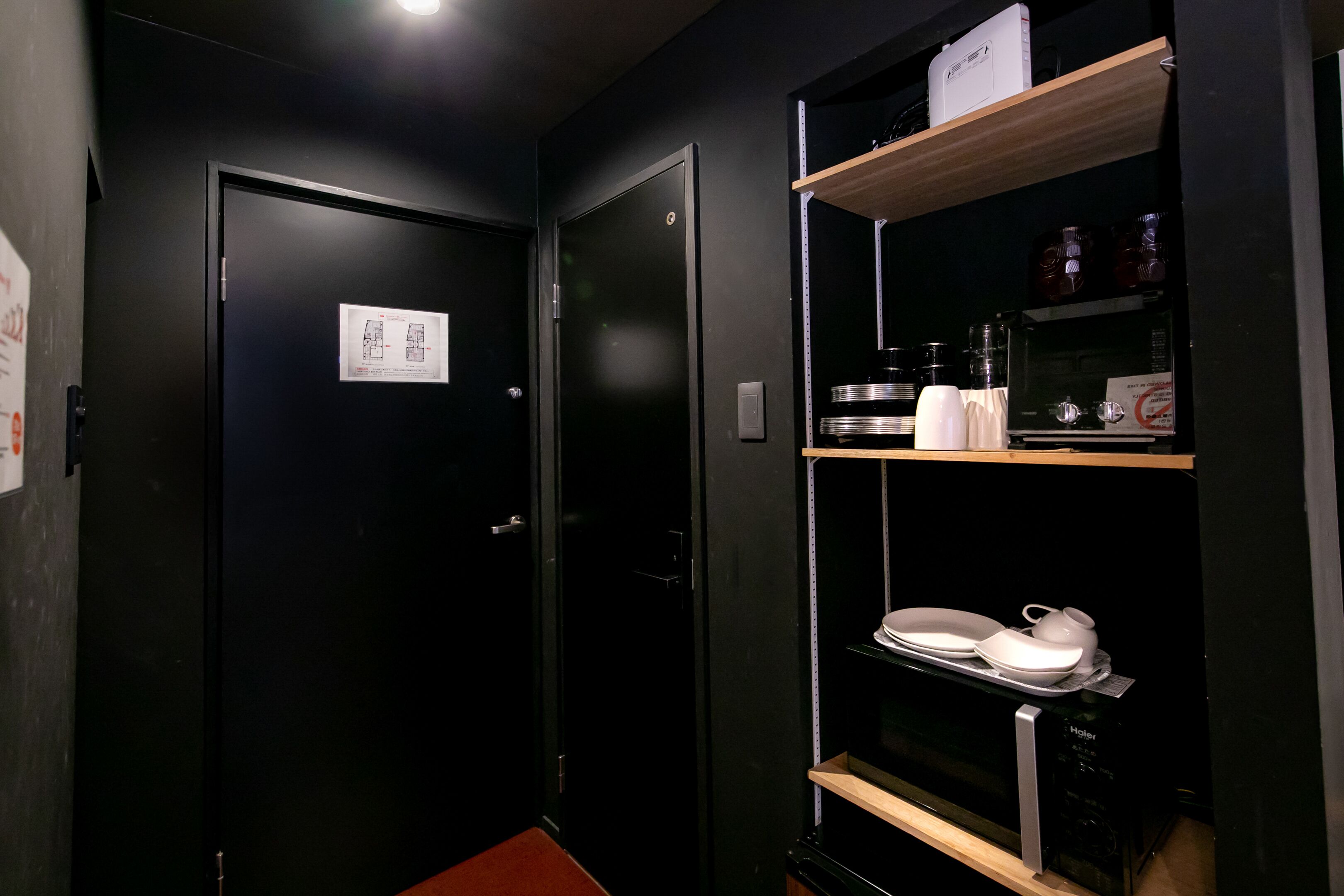 Un couloir étroit avec une porte noire, une étagère en bois contenant des ustensiles de cuisine et un four à micro-ondes.