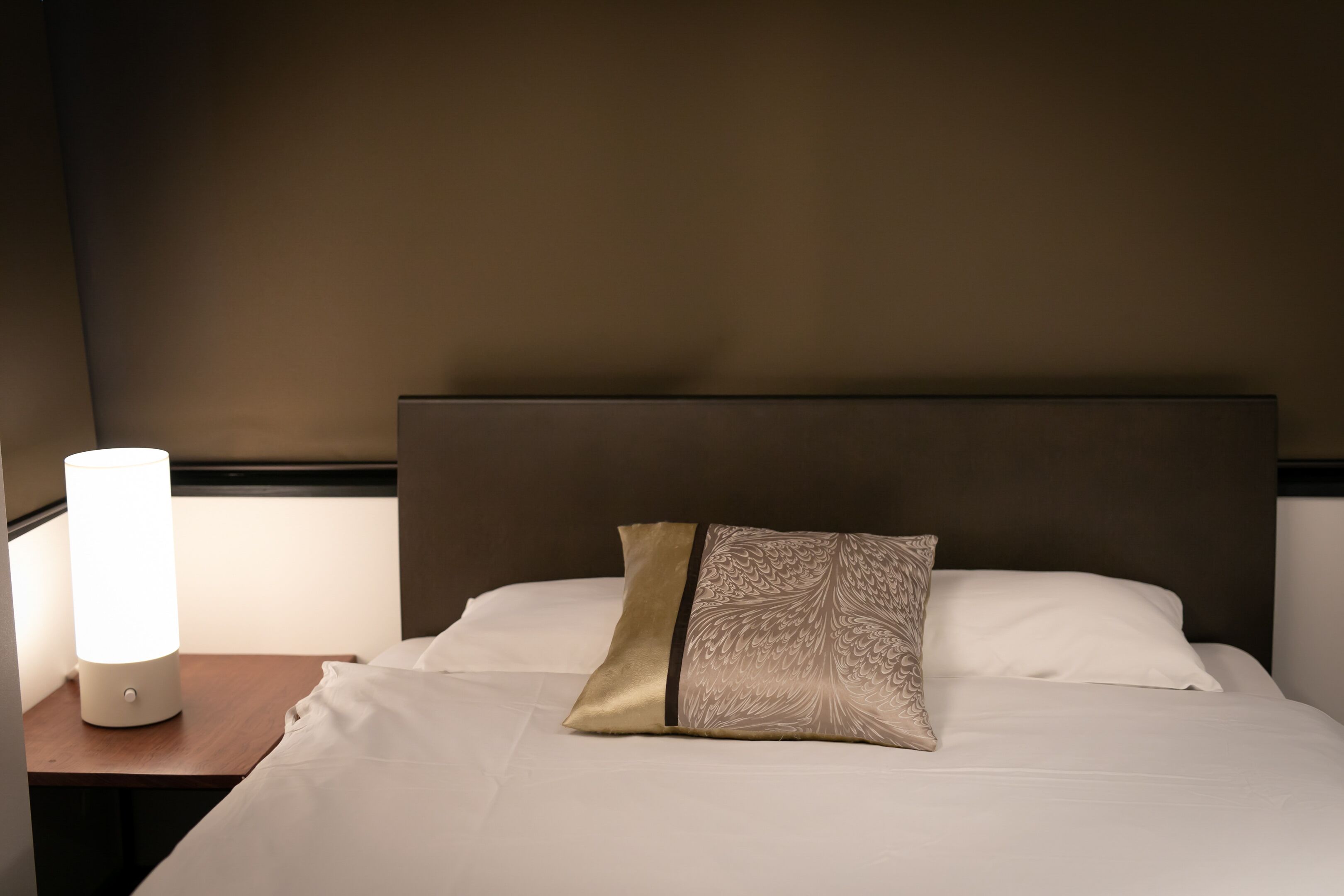 Un lit bien fait, agrémenté d’un coussin décoratif et d’une lampe de chevet posée sur une table de nuit en bois.