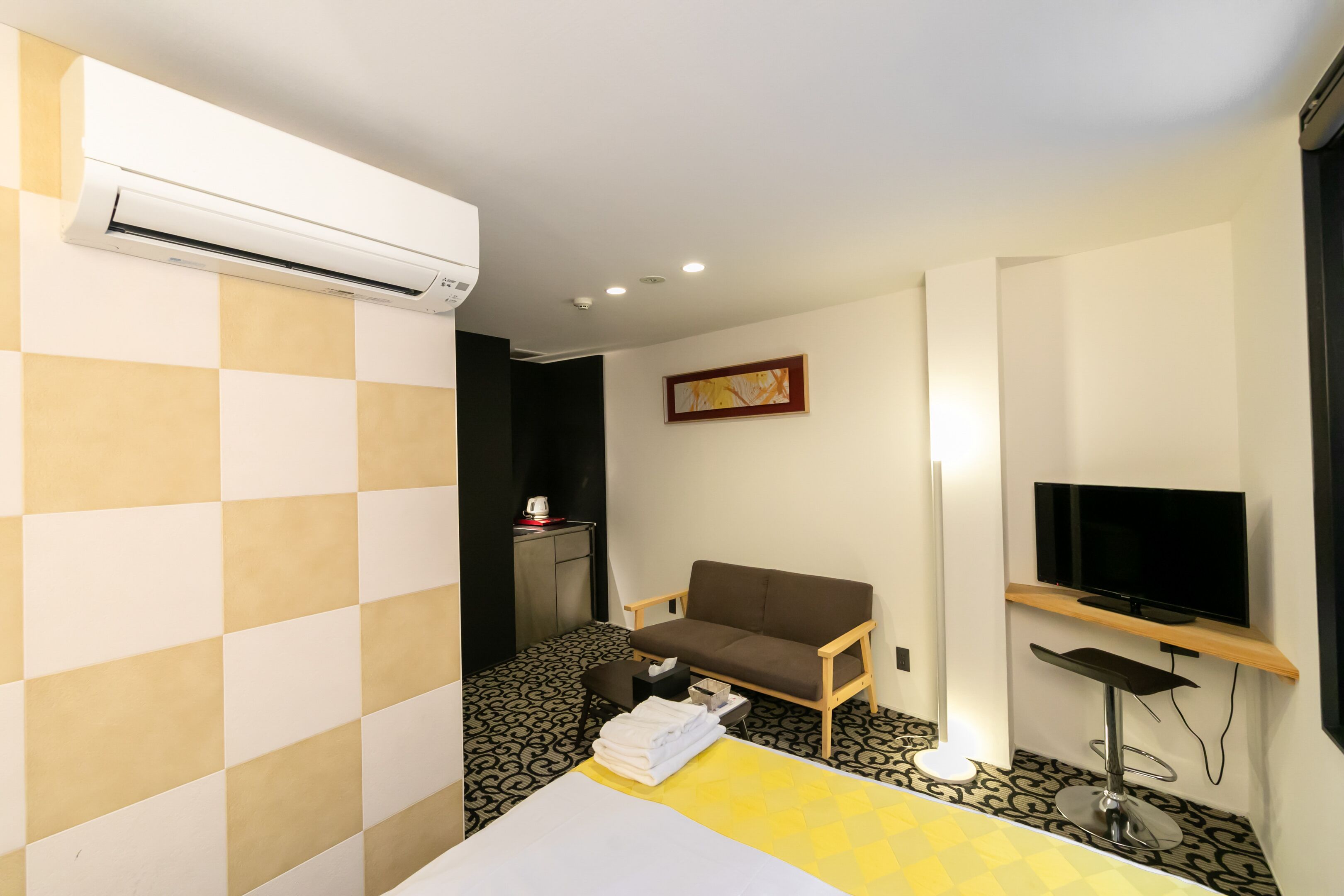 A hotel room with a bed, a sofa, a TV, an air conditioner, a desk, and a small table with a lamp.
