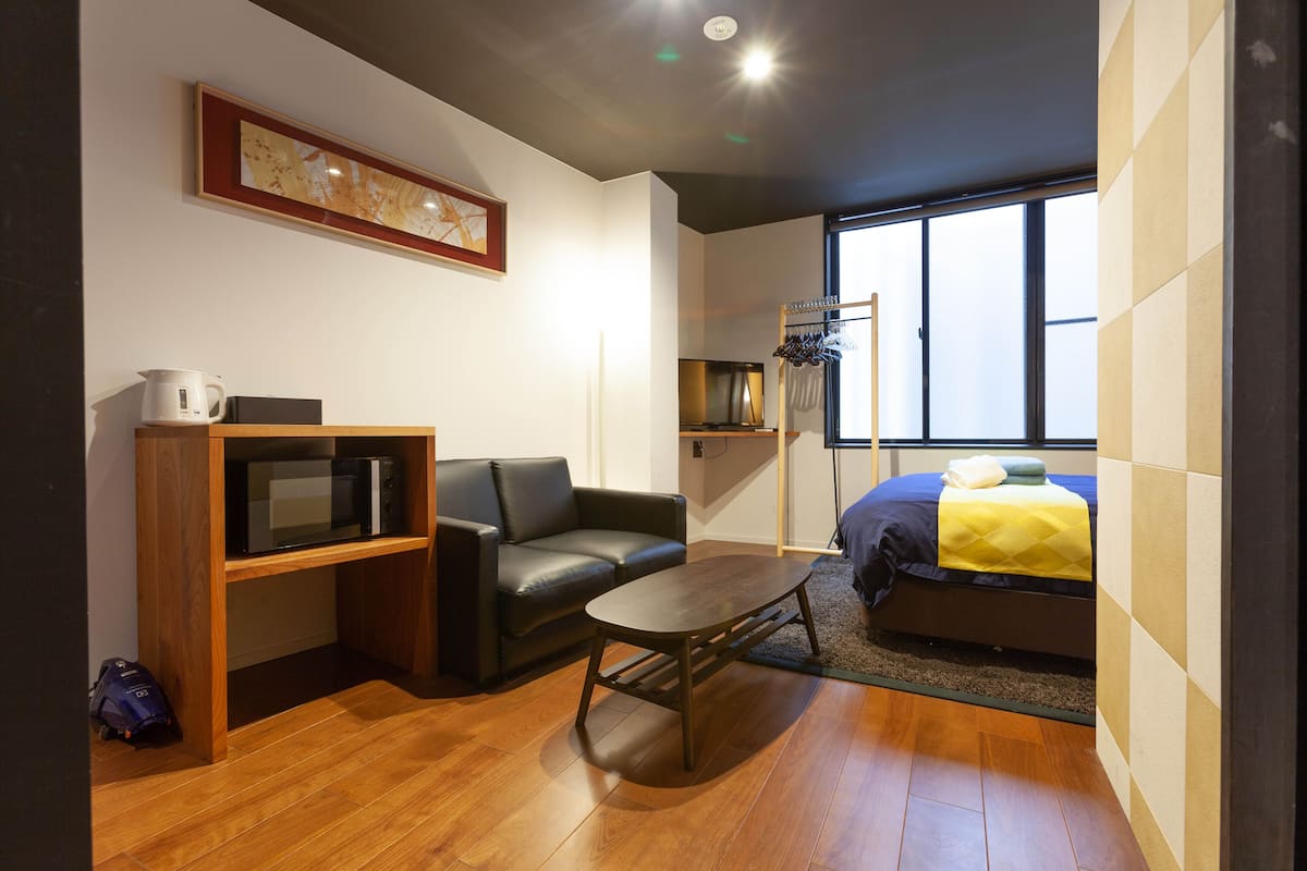 Ein gemütlicher Raum mit Holzboden, einer schwarzen Ledersofa, einem kleinen Tisch, einem Bett mit gelber Decke, einem Fernseher auf einer Holzablage und einem gerahmten Bild an der Wand.