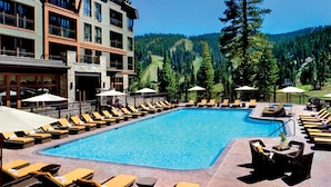 Un espace piscine doté de chaises longues et de parasols, entouré d’un immeuble à plusieurs étages et avec une chaîne de montagnes en arrière-plan.