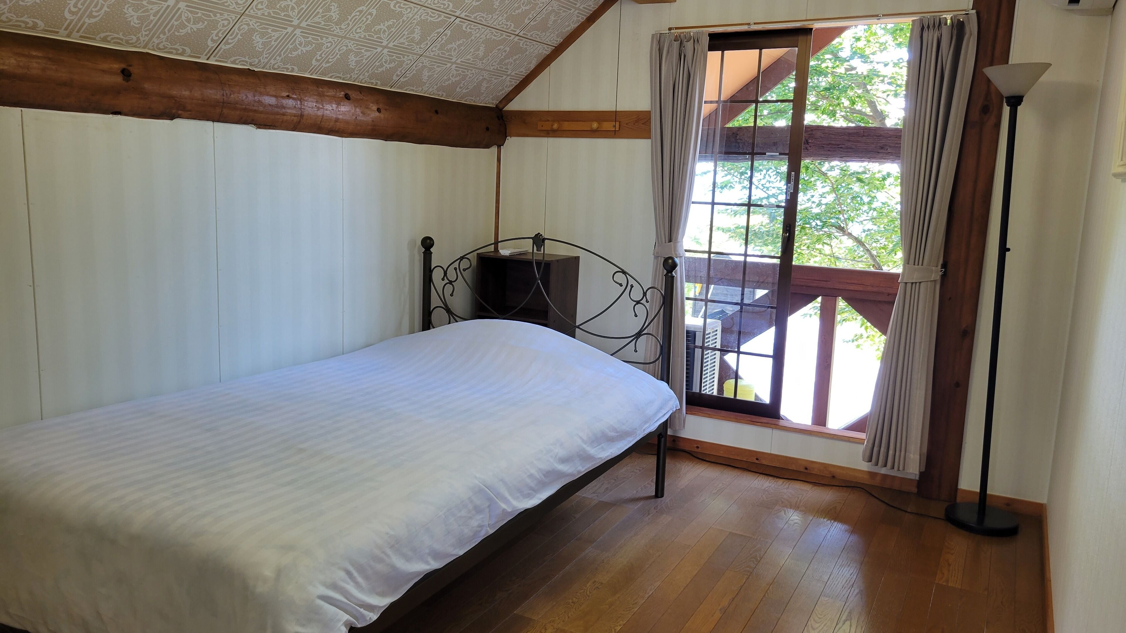 Un dormitorio con techo de madera, cama con colchón blanco, cabecero de metal, ventana con cortinas y una lámpara de pie.