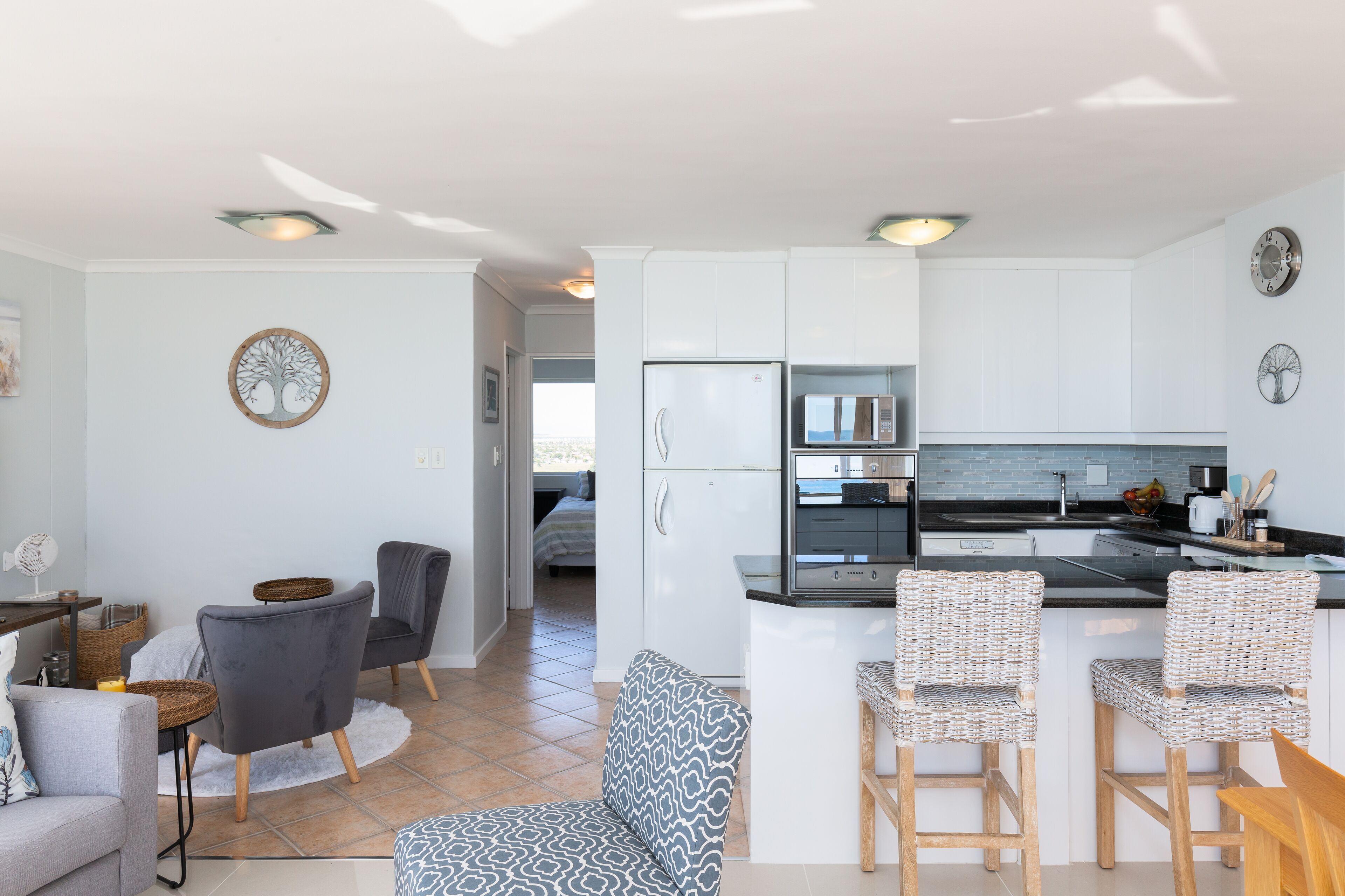 A modern kitchen with white cabinets, a built-in microwave, and a refrigerator. There are wicker chairs and a small dining table.