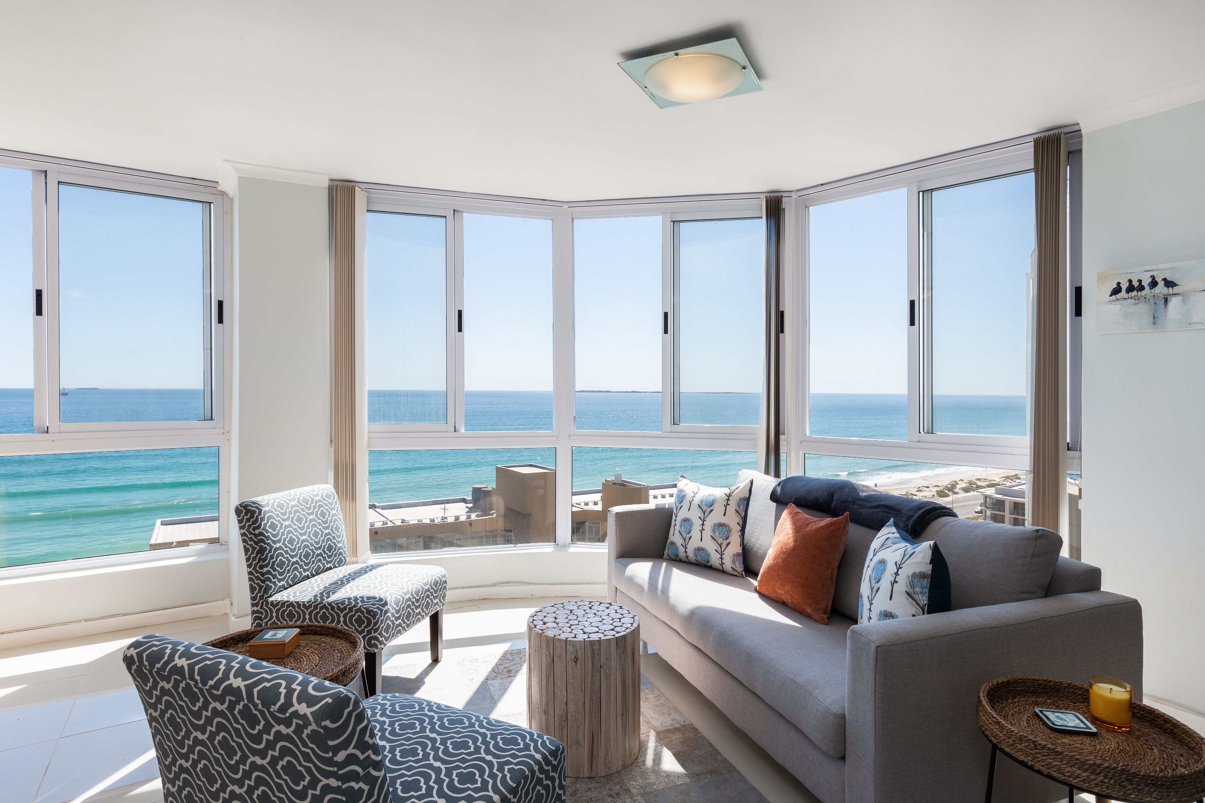 A modern living room with a large bay window overlooking the sea, a grey sofa with patterned cushions, and a small round table with a candle.