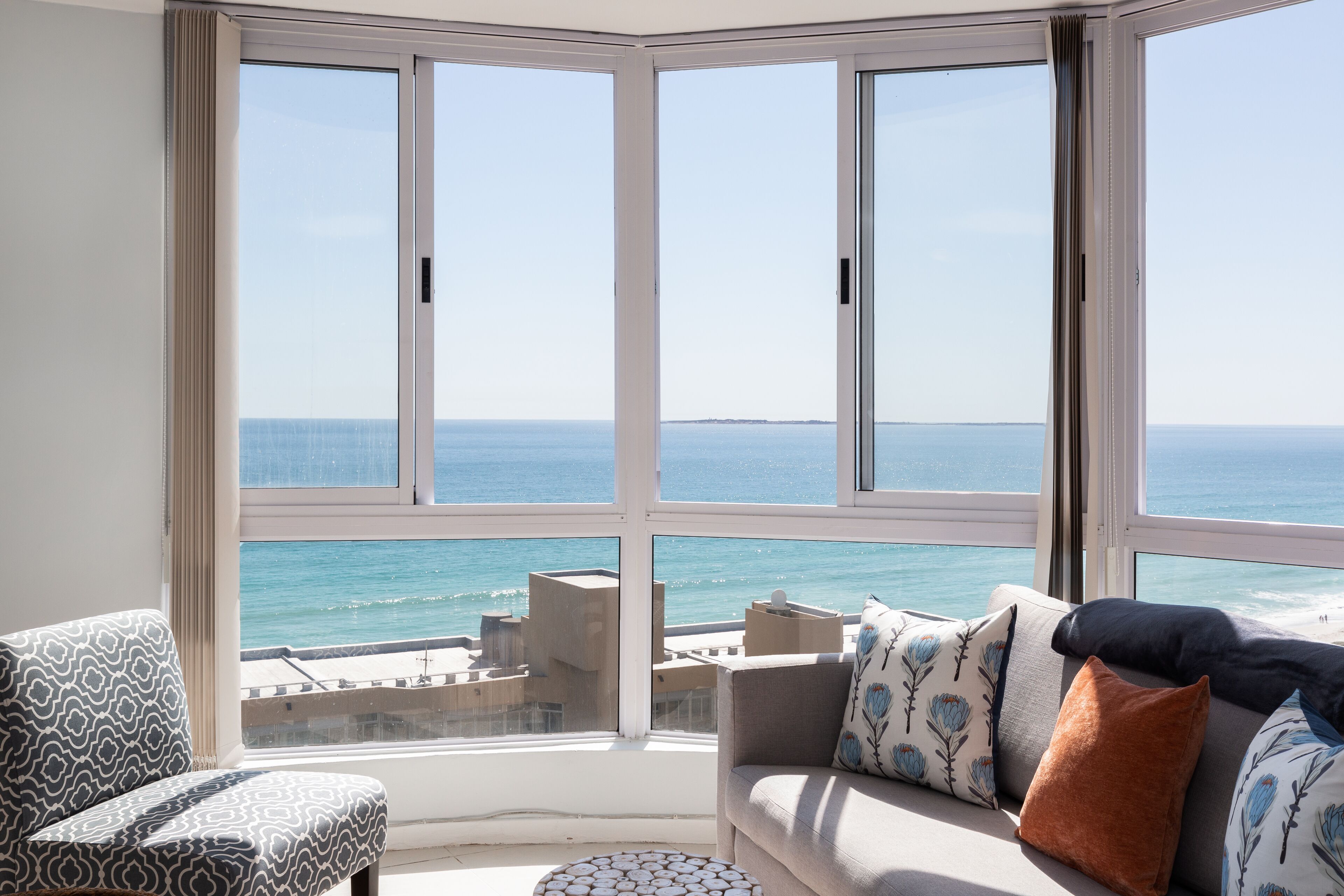 A room with a large bay window overlooking the sea, a sofa with patterned cushions, and a patterned armchair.