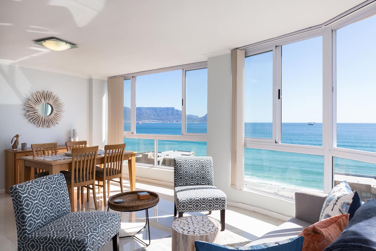 A spacious room with a large window overlooking the sea, a dining area with wooden furniture, and patterned seating.