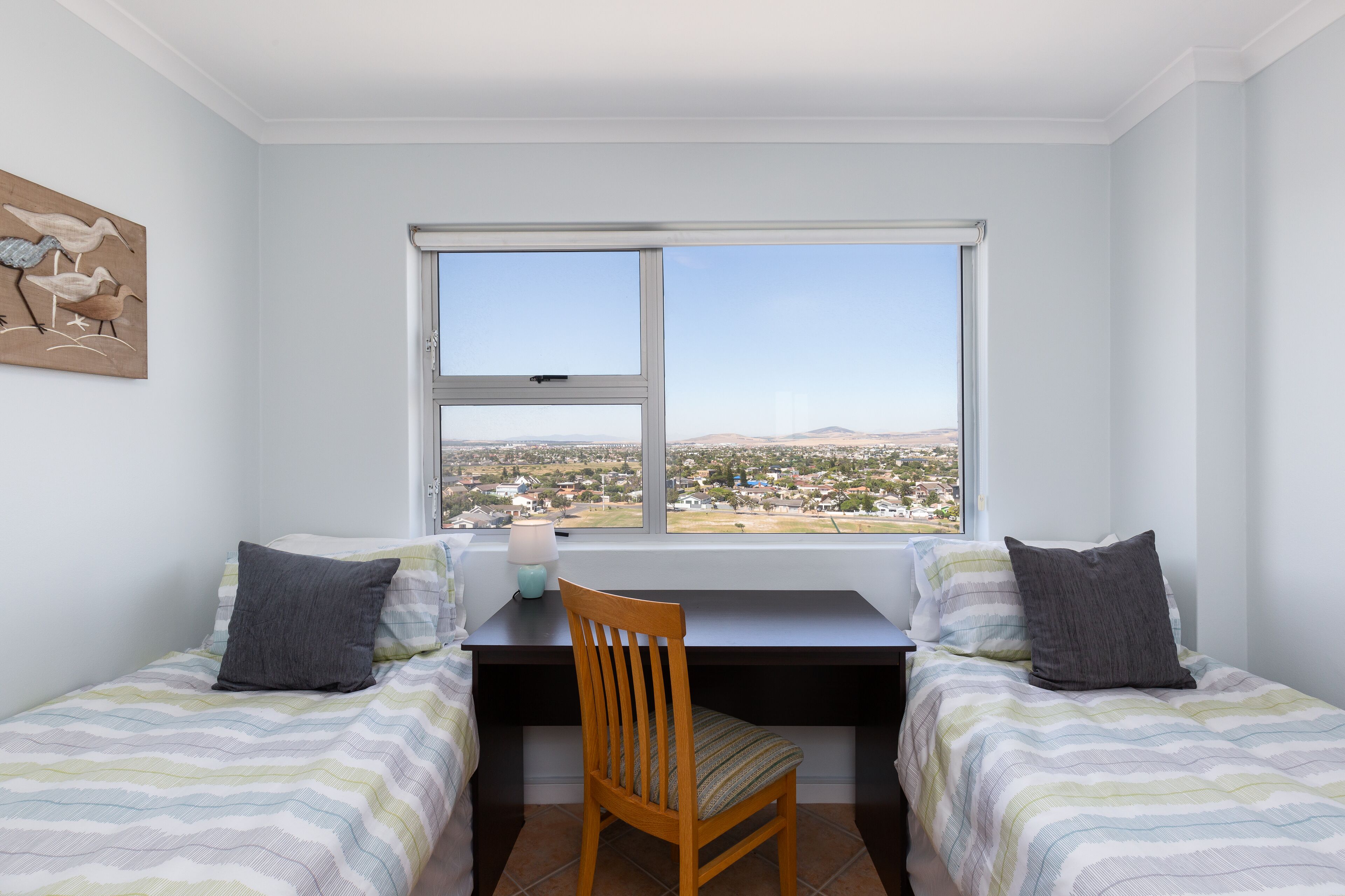Una camera da letto con due letti singoli, una scrivania e una sedia. Si vede una finestra con vista su una città e montagne.