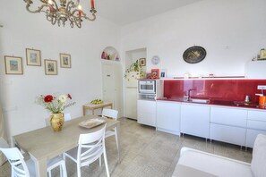 Una cocina moderna con armarios blancos, encimera roja y una araña. En la pared hay cuadros enmarcados y un jarrón con flores sobre la mesa.