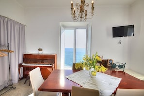 Zona pranzo con un pianoforte, un tavolo con un vaso di fiori, un lampadario e una TV appesa al muro.