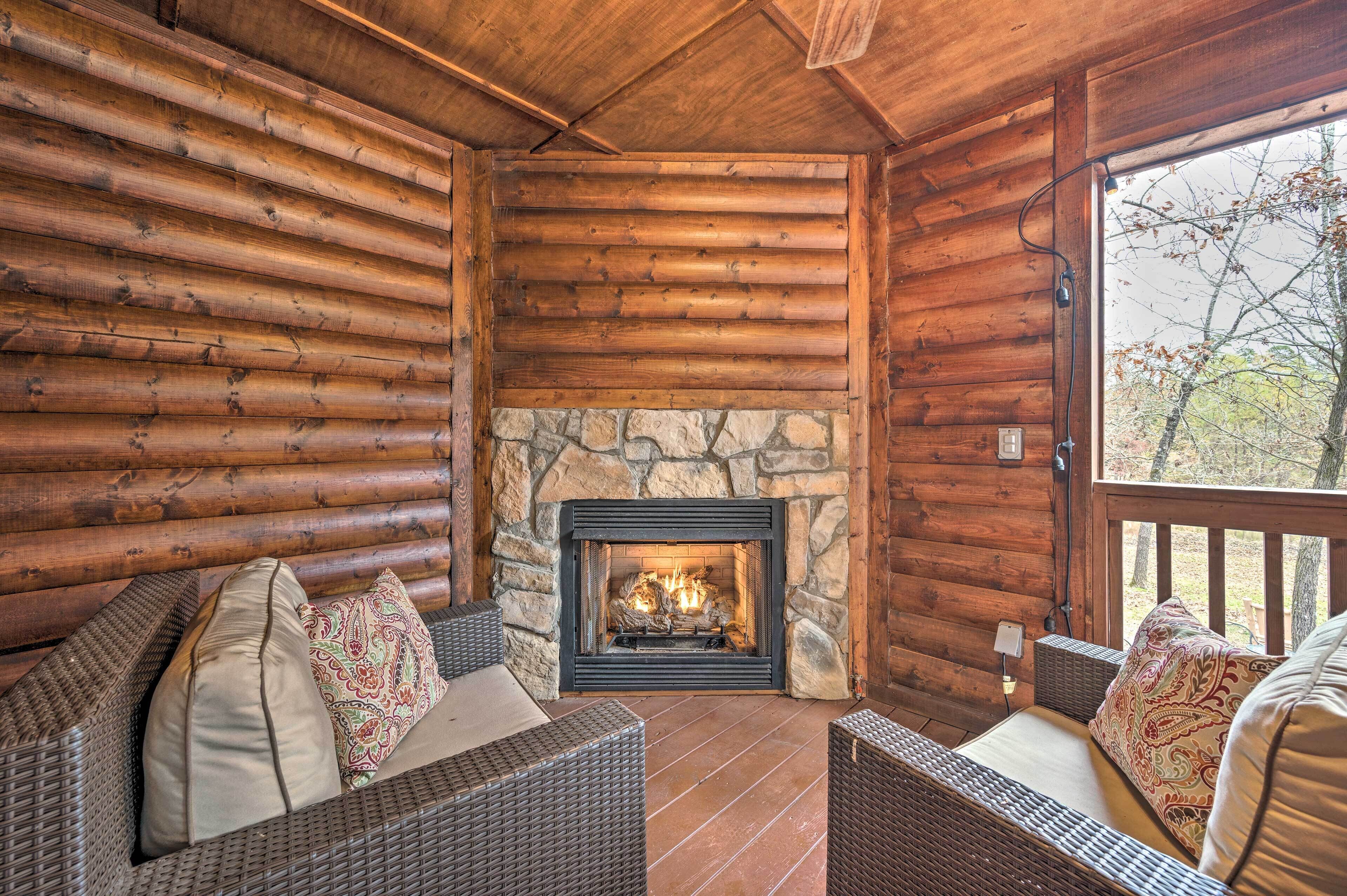 A log cabin interior with a stone fireplace, wicker furniture, and a view of trees outside.