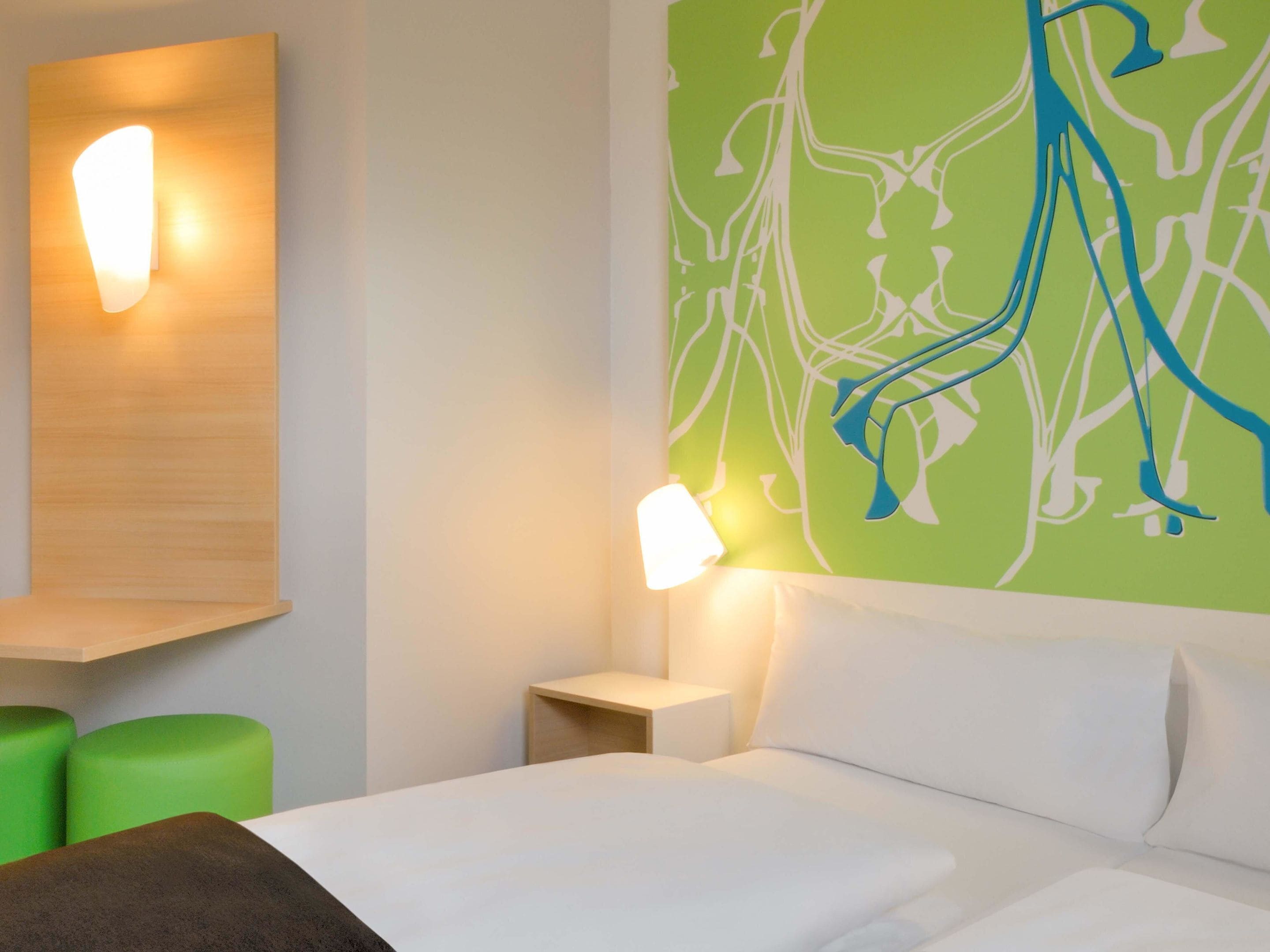 Ein Hotelzimmer mit einem Bett, grünen Polsterstühlen, einer Wandleuchte und einer Wand mit abstrakten Mustern.