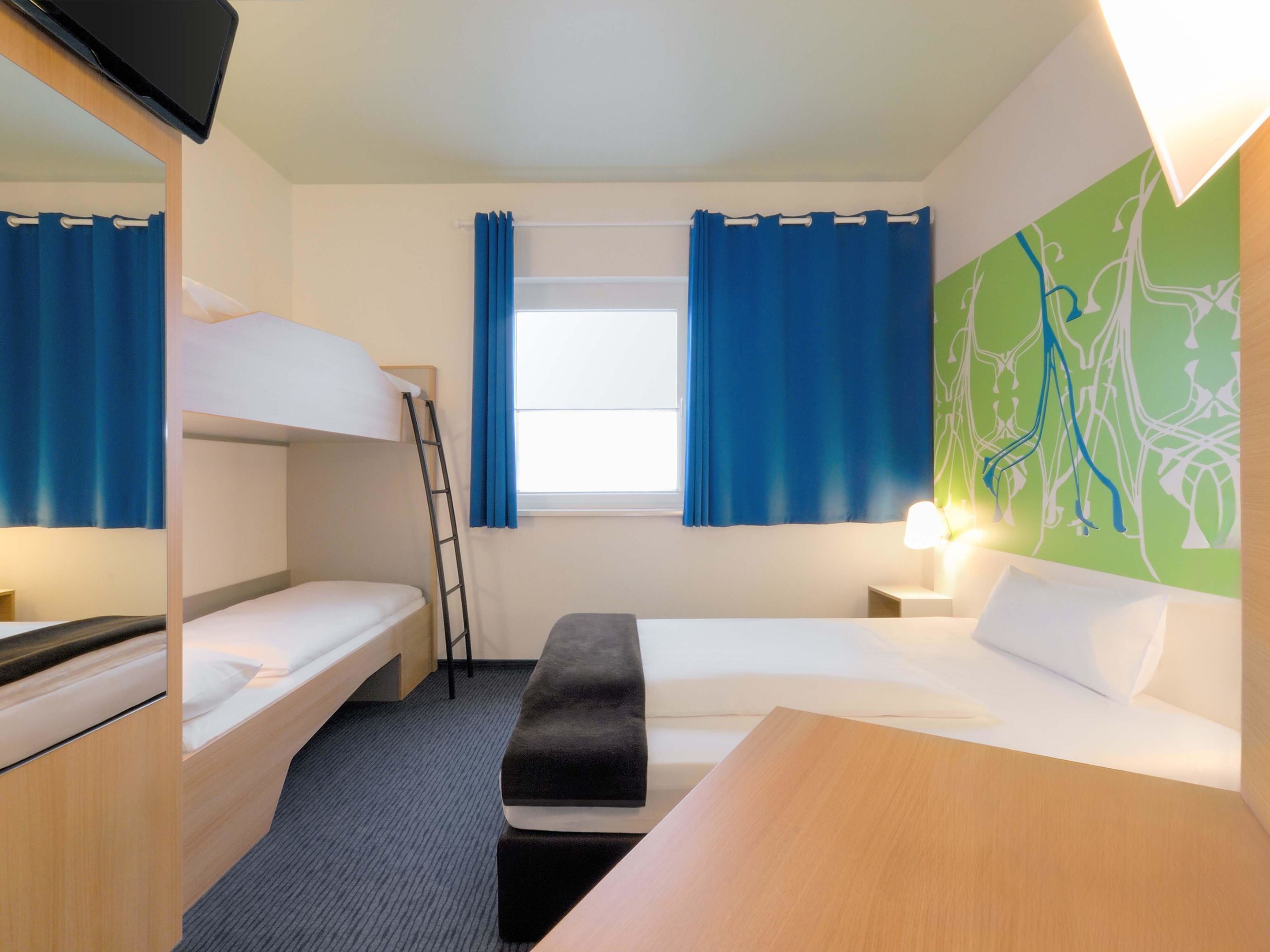 A hotel room with bunk beds, a desk, a chair, a small table, a lamp, and a window with curtains.