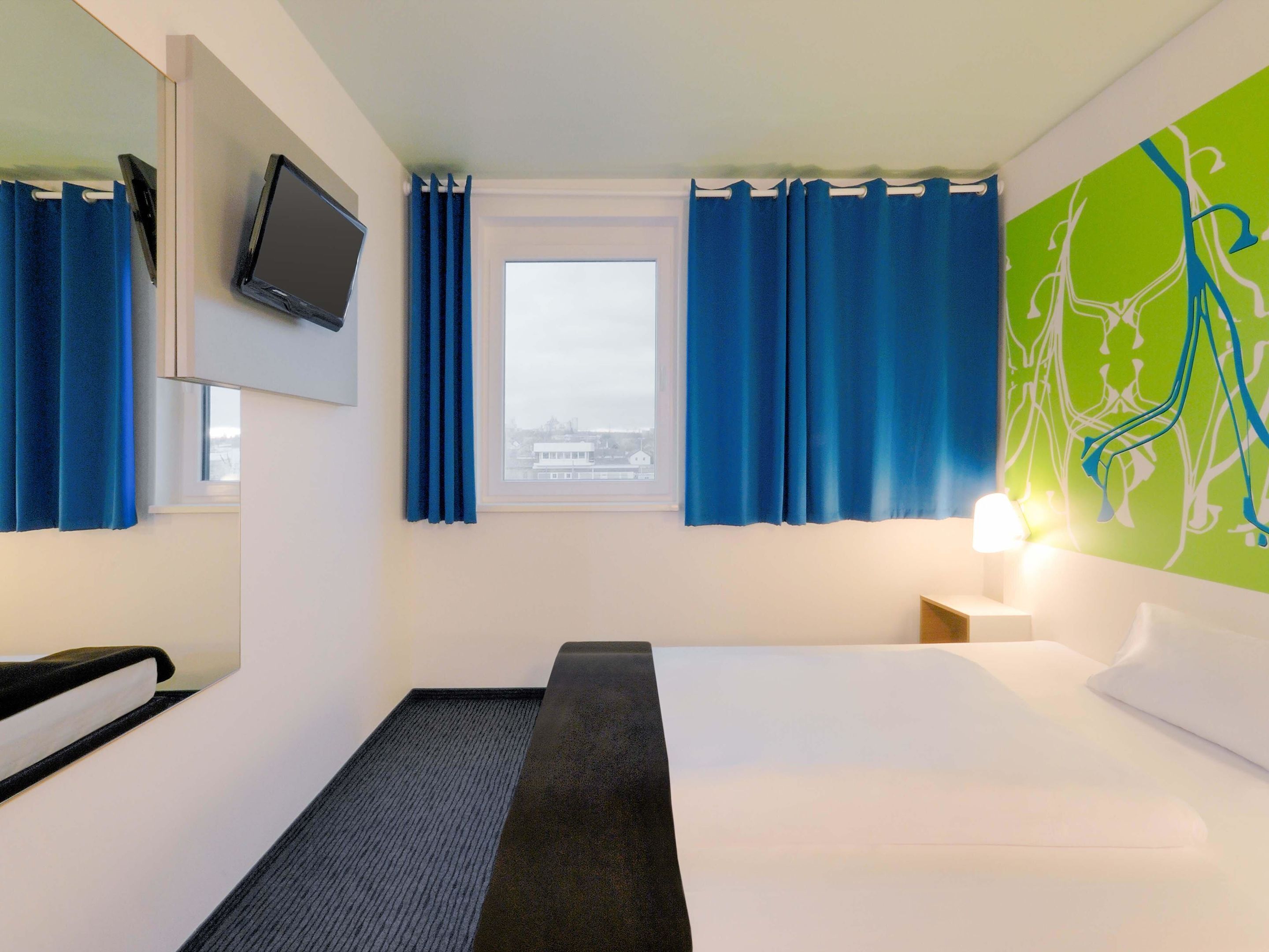 Une chambre d’hôtel avec un lit, un bureau, une chaise, une télévision et une fenêtre avec des rideaux.
