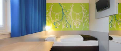 A hotel room with a bed, a desk, a chair, a TV mounted on the wall, and a window with curtains.
