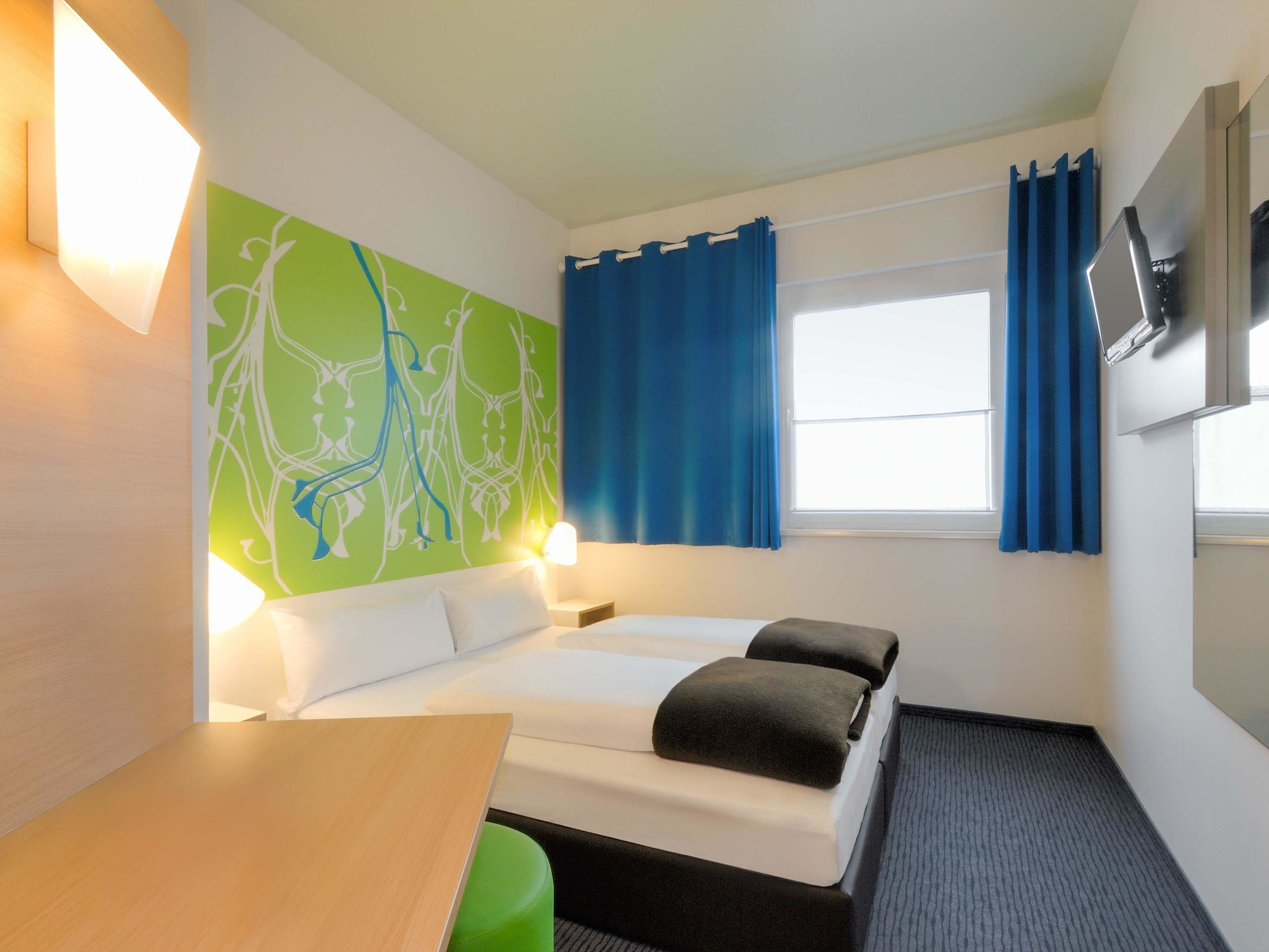 A hotel room with a bed, a desk, a green wall with a white abstract design, a window with blue curtains, and a flat-screen TV mounted on the wall.