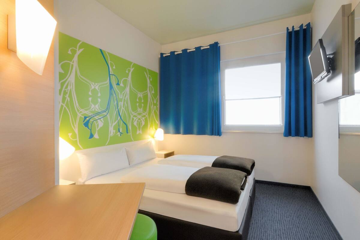 A hotel room with a bed, a desk, a green wall with a white abstract design, a window with blue curtains, and a flat-screen TV mounted on the wall.