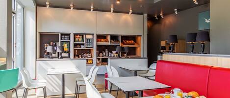 Un café moderno con mesas y sillas blancas, un rincón con bancos rojos, y una amplia variedad de comida y bebidas.
