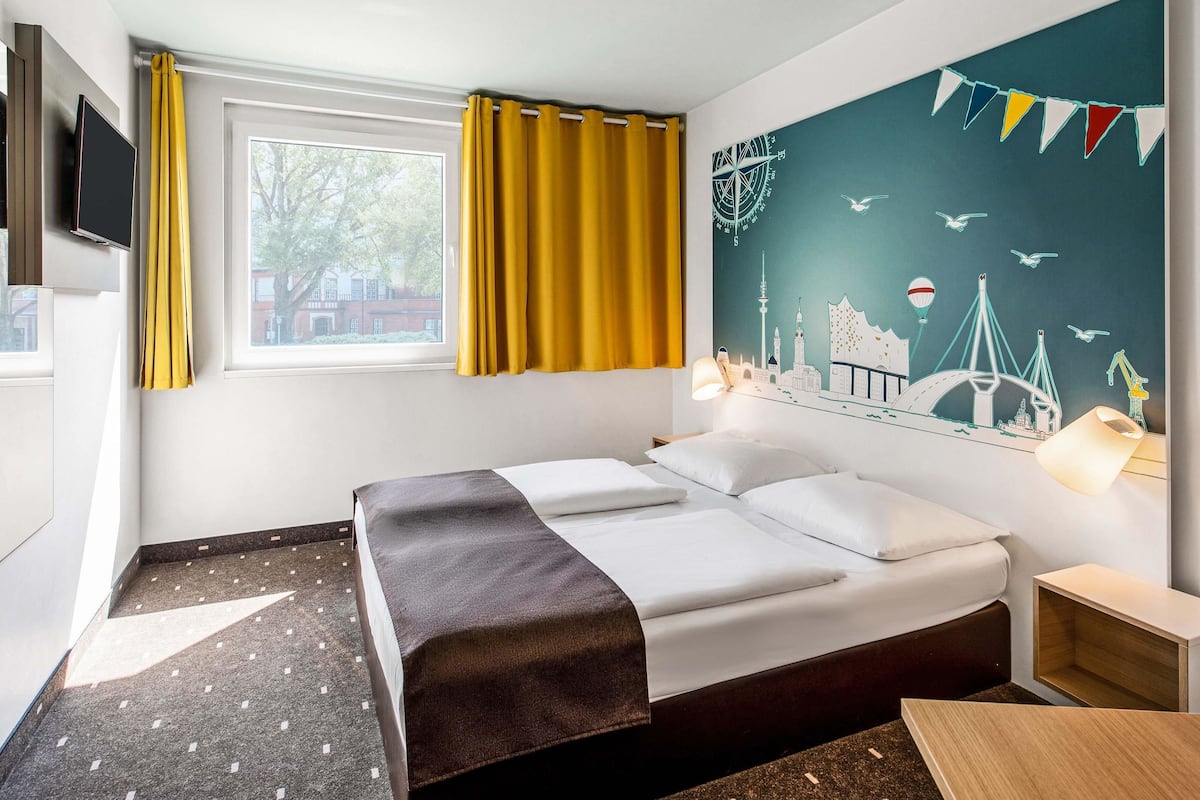A hotel room with a bed, a window with curtains, a wall-mounted TV, and a cityscape mural.