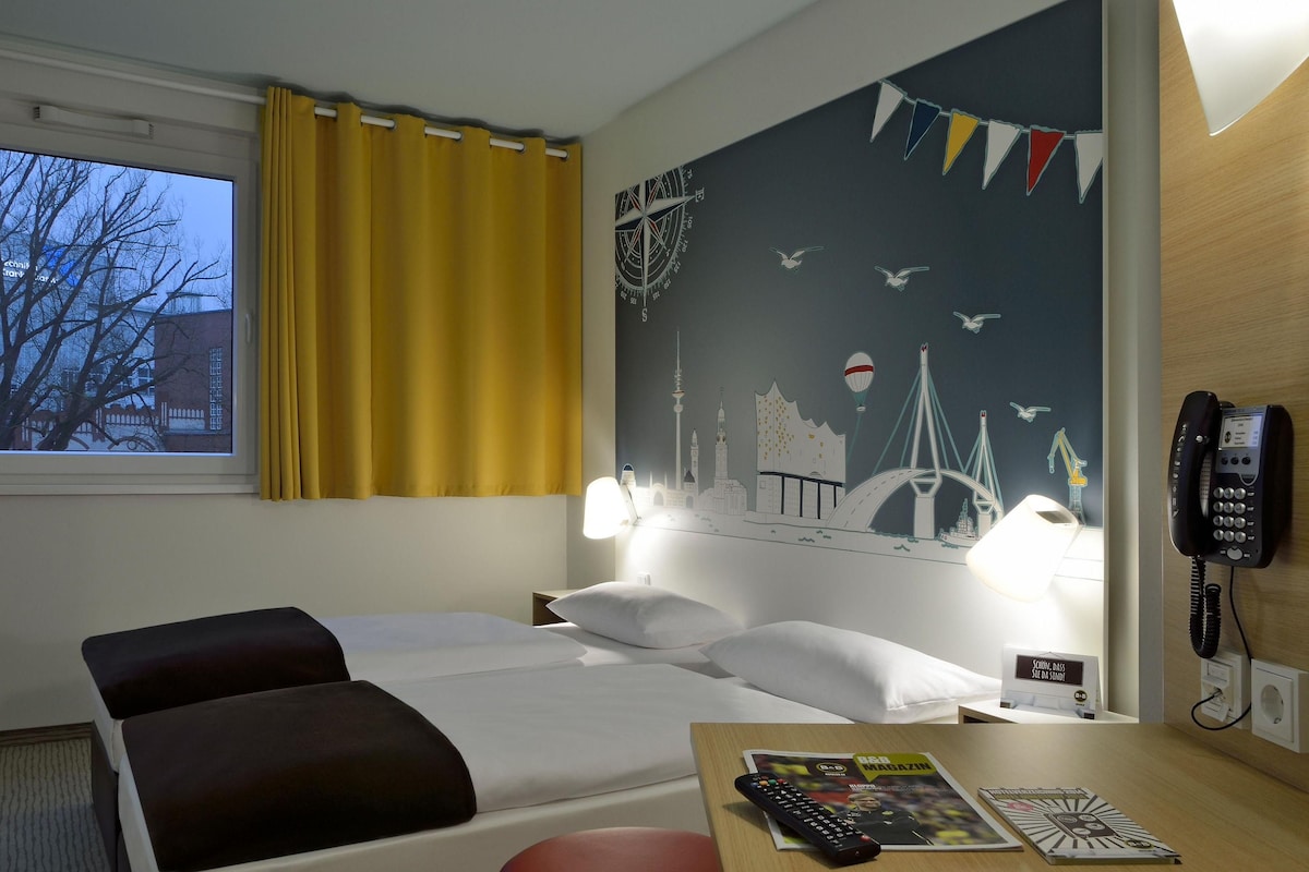 A hotel room with a bed, two pillows, a nightstand with a lamp, a wall clock, a phone, and a window with curtains.