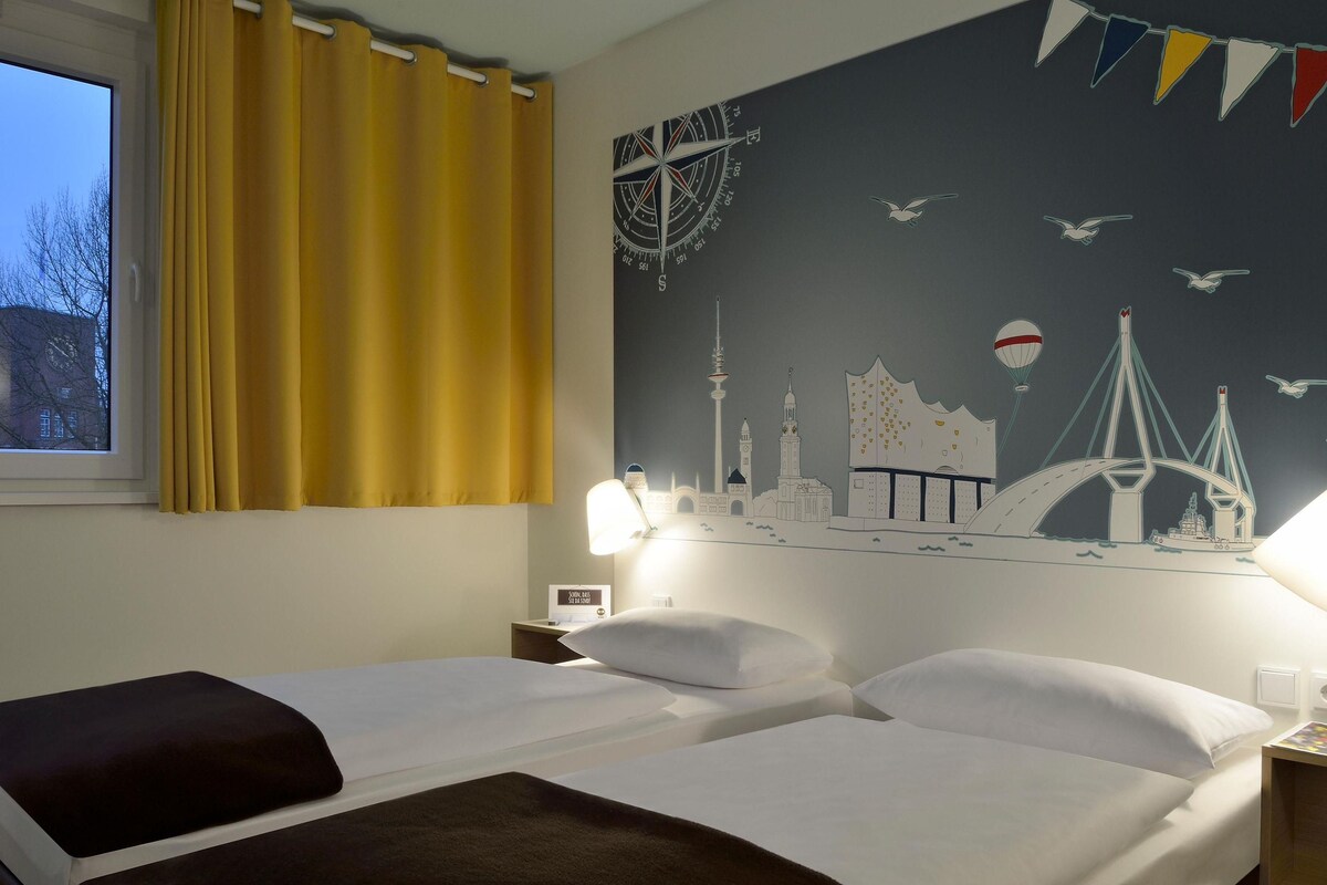 A hotel room with two beds, a window with yellow curtains, a wall mural of a cityscape, and a nightstand with a lamp.