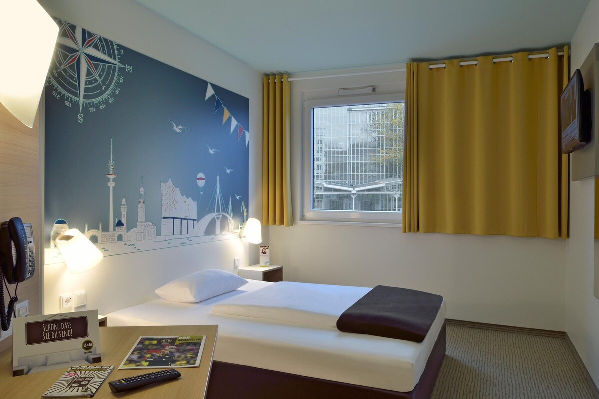 A hotel room with a bed, bedside lamps, a wall clock, a window with curtains, and a wall with a compass and cityscape mural.
