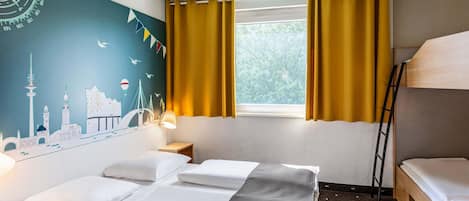 A hotel room with a bed, a desk, a lamp, a window with curtains, and a wall mural.