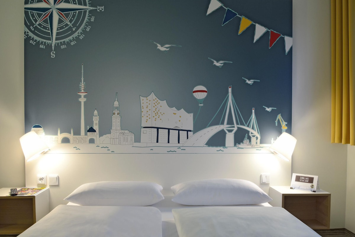 A hotel room with a bed, two bedside tables, a wall mural of a city skyline, and a compass.