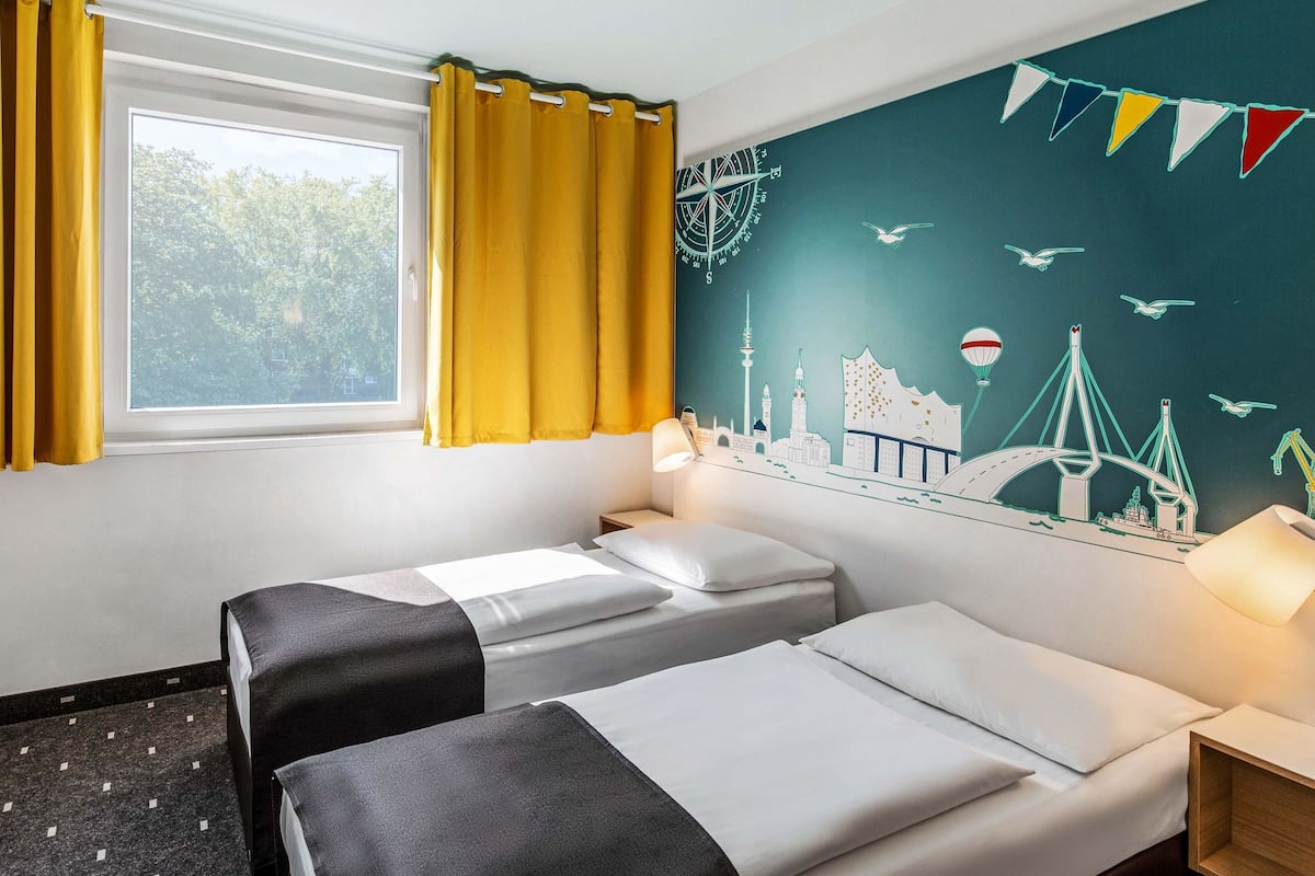 A hotel room with two beds, a window with yellow curtains, a wall mural of a cityscape, and a nightstand with a lamp.