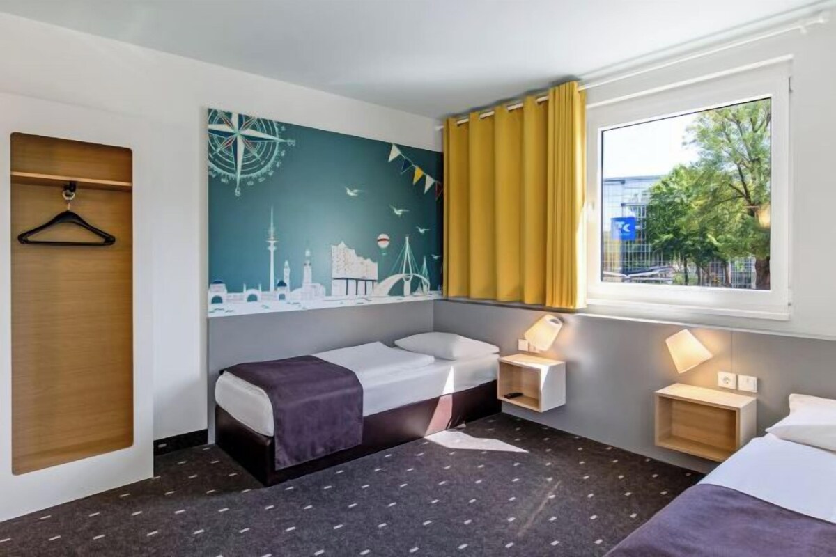 A hotel room with a bed, a bedside table with a lamp, a window with curtains, and a wall with a cityscape mural.