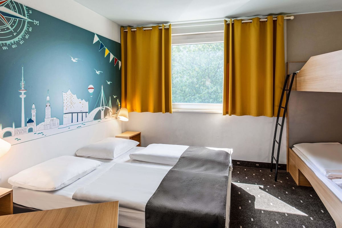 A hotel room with a bed, a desk, a lamp, a window with curtains, and a wall mural.