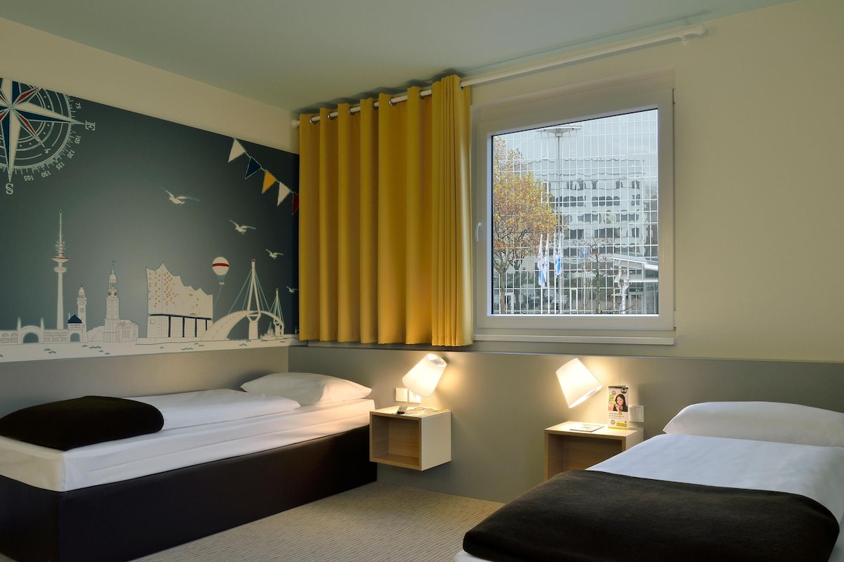 A hotel room with two beds, a cityscape mural, a window with yellow curtains, and a view of buildings outside.